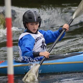 Junge im blauen Kajak paddelt konzentriert durch einen Sportkanal, mit Helm und Schwimmweste ausgestattet.