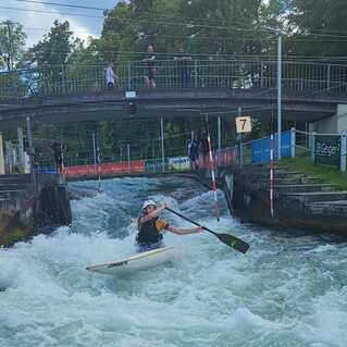 Kajakfahrer paddelt durch reißendes Wasser unter einer Brücke, Zuschauer beobachten vom Geländer.