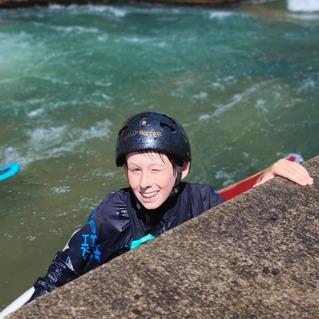 Junger Wassersportler mit Helm lächelt und winkt, während er an einer Steinmauer in einer Wasserfläche steht.