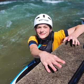 Junger Mann in einem Kanu mit Helm greift nach einem Steg an einem künstlichen Wildwasserkanal.