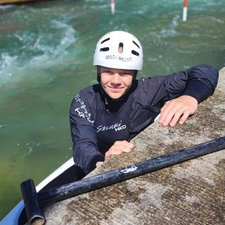 Junger Mann mit Helm in einem Kanu, der sich an einem Steg über einem Wasserkanal festhält.