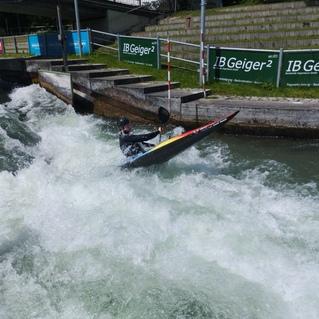 Kajakfahrer manövriert durch aufgewühltes Wasser in einem Wildwasserkanal, umgeben von Werbebannern und neuer grüner Vegetation.