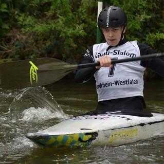Junger Kanute in einem weißen Shirt paddelt auf einem Fluss, spritzendes Wasser um ihn herum.