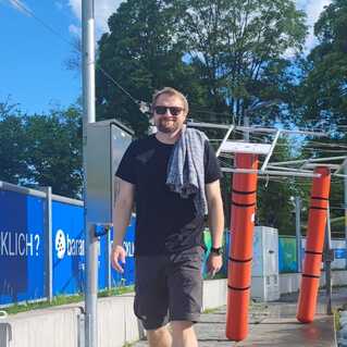 Mann mit Handtuch über der Schulter geht auf einem Gehweg entlang einer Kanalwasserbahn mit roten Markierungspfosten.