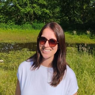 Frau mit Sonnenbrille lächelt vor einer grünen Landschaft mit Bäumen und einem kleinen Gewässer.