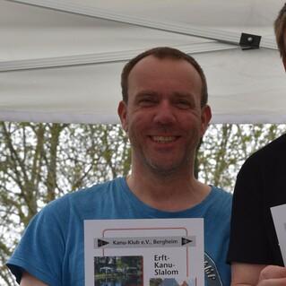 Mann hält Urkunde für den 2. Platz beim Erft-Kanu-Slalom, umgeben von Bäumen und einem Zelt.