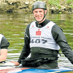 Sportler im Kanu mit Helm und Schwimmweste, lächelt am Ufer eines Gewässers.