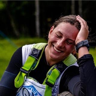 Lächelnde Person mit langen Haaren, die auf einem Tisch lehnt, umgeben von grüner Natur.