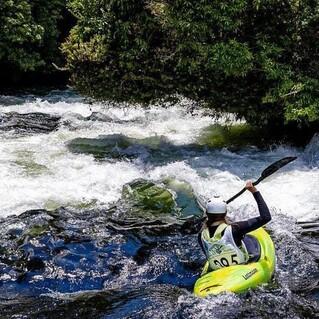 Kanufahrer paddelt auf einem wild fließenden Fluss mit schäumendem Wasser und grünen Bäumen im Hintergrund.