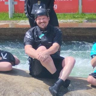 Fünf Personen in Neoprenanzügen sitzen auf einem großen Felsen im Wasser; im Hintergrund sind Zuschauertribünen sichtbar.