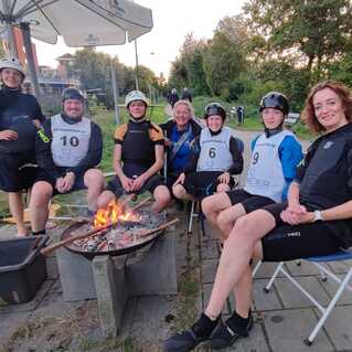 Gruppe von sieben Personen in Sportkleidung, sitzend um ein Lagerfeuer an einem Gewässer, mit Bäumen im Hintergrund.
