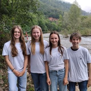 Vier Jugendliche in weißen T-Shirts stehen an einem Fluss, umgeben von Bäumen und Hügeln im Hintergrund.