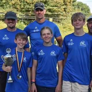 Sechs Personen in blauen T-Shirts stehen am Ufer eines Gewässers, ein Junge hält einen Pokal und eine Medaille.