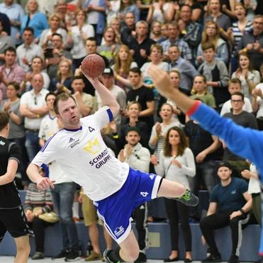 Handballspieler in weißem Trikot wirft einen Ball über einen gegnerischen Spieler, im Hintergrund jubelndes Publikum.