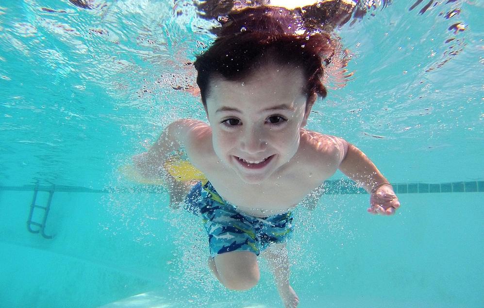 Junge schwimmt unter Wasser mit lächelndem Gesicht und bunten Schwimmshorts in einem Pool. Blasen entstehen um ihn herum.