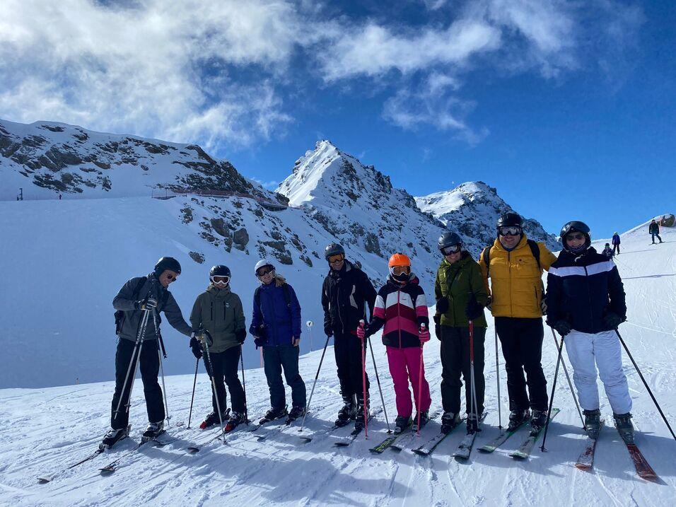 Eine Gruppe von acht Skifahrern posiert im Schnee vor verschneiten Bergen bei blauem Himmel.