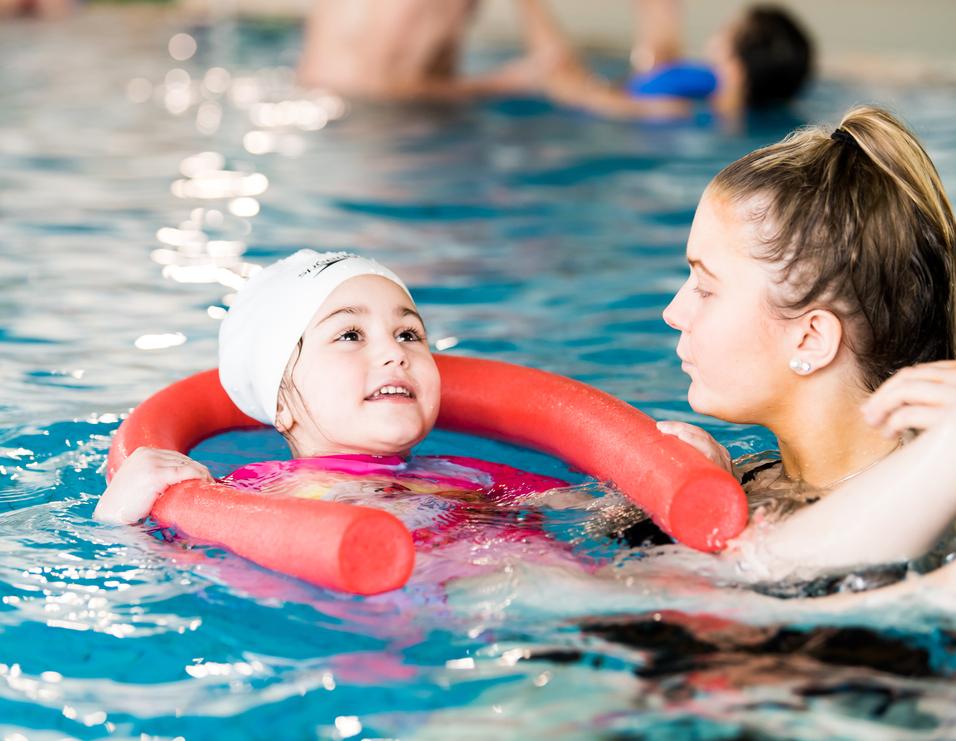Ein Mädchen mit Schwimmring lächelt in einem Schwimmbecken, während eine Schwimmtrainerin neben ihr steht.
