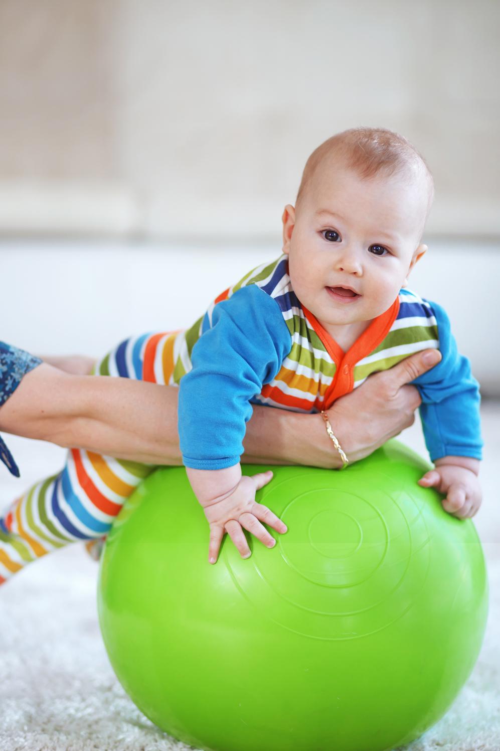 Baby in bunten Strampelanzug liegt auf einem grünen Gymnastikball, von einer Hand unterstützt.